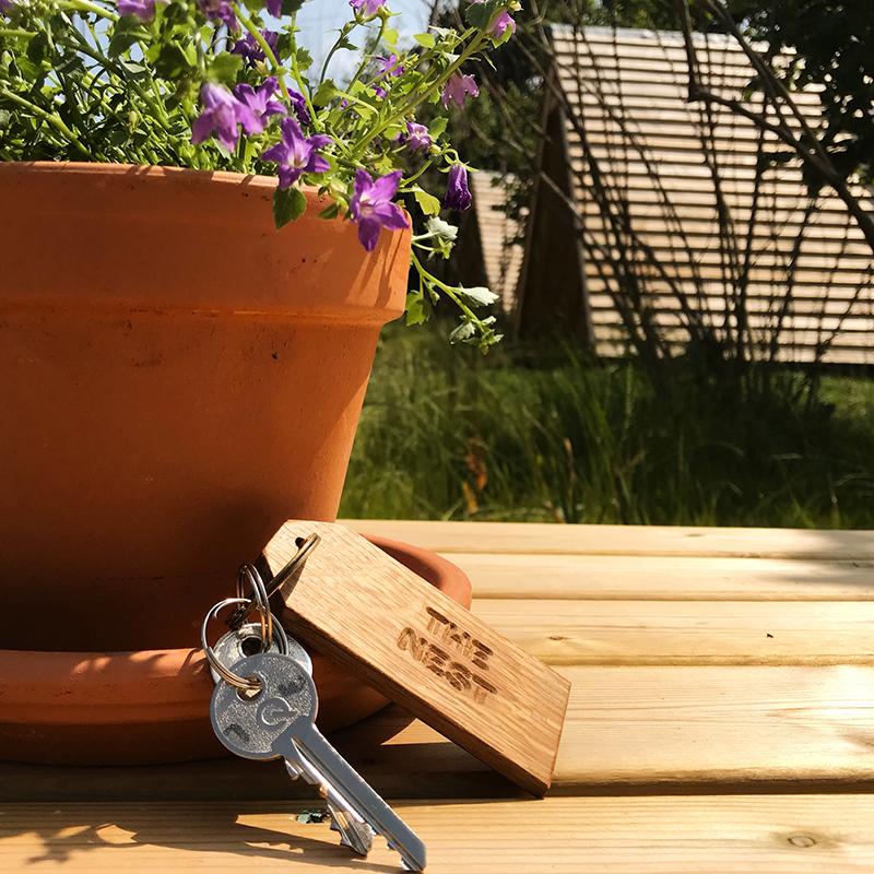 Door key with wooden fob against a flower pot with purple flowers on a picnic bench with Hen's Dens in the background